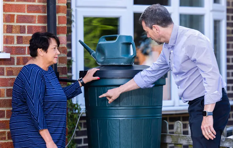 Two people stood in a garden next to a water butt with a watering can on the top 