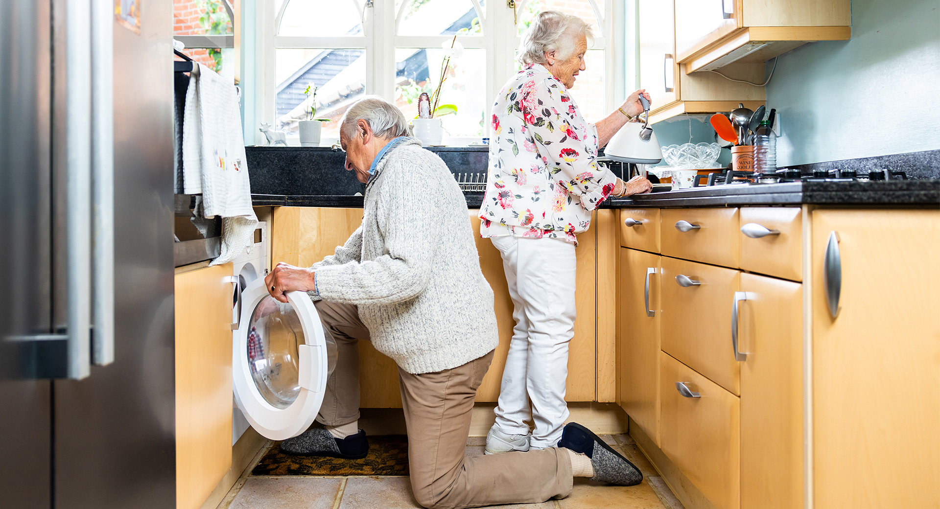 An elderly couple in their kitchen, one prepares food while the other puts a towel into the washing machine
                        